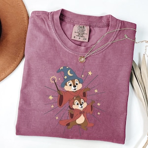May include: A mauve Comfort Colors t-shirt featuring a cartoon graphic of two chipmunks dressed as wizards, with a wizard hat and cape. The chipmunks are surrounded by starbursts and stars. A gold necklace with a pendant is on the shirt.