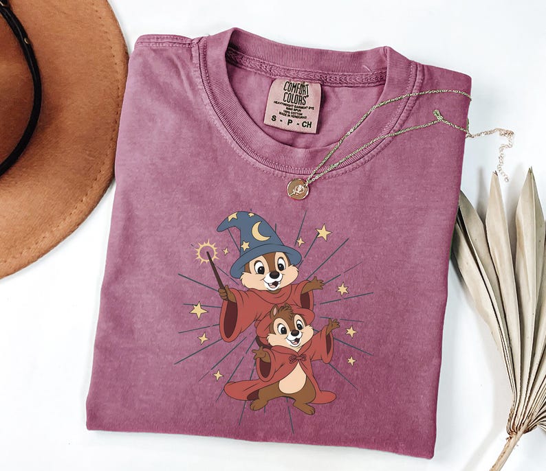 May include: A mauve Comfort Colors t-shirt featuring a cartoon graphic of two chipmunks dressed as wizards, with a wizard hat and cape. The chipmunks are surrounded by starbursts and stars. A gold necklace with a pendant is on the shirt.