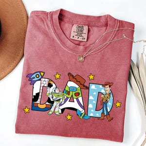 May include: A red t-shirt with the word "DAD" in a colourful design featuring characters from the movie Toy Story. The shirt has a crew neck and a small tag at the neckline. A gold necklace is draped across the shirt.