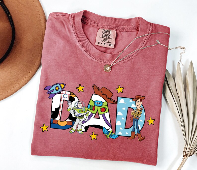 May include: A red t-shirt with the word "DAD" in a colourful design featuring characters from the movie Toy Story. The shirt has a crew neck and a small tag at the neckline. A gold necklace is draped across the shirt.