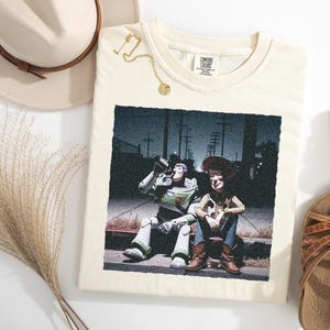 May include: A cream-colored t-shirt featuring a graphic print of Buzz Lightyear and Woody sitting on a curb at night. The shirt has a Comfort Colors label at the neckline. The design shows the two characters in a dark, urban setting with streetlights.