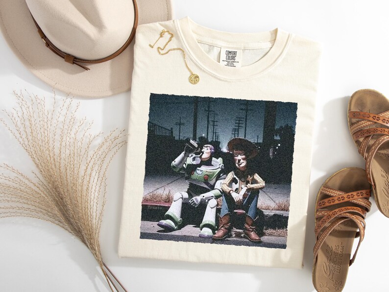 May include: A cream-colored t-shirt featuring a graphic print of Buzz Lightyear and Woody sitting on a curb at night. The shirt has a Comfort Colors label at the neckline. The design shows the two characters in a dark, urban setting with streetlights.
