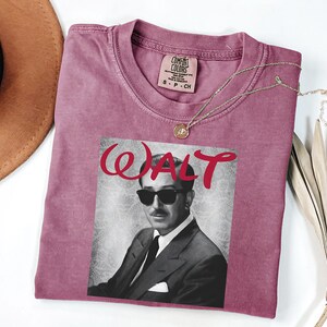 Op de afbeelding: Een stoffig roze t-shirt met een zwart-wit afbeelding van een man met een zonnebril en het woord "WALT" in rode letters. Het shirt heeft een ronde hals en een klein label bij de halslijn. Een gouden ketting met een hanger is over het shirt gedrapeerd.