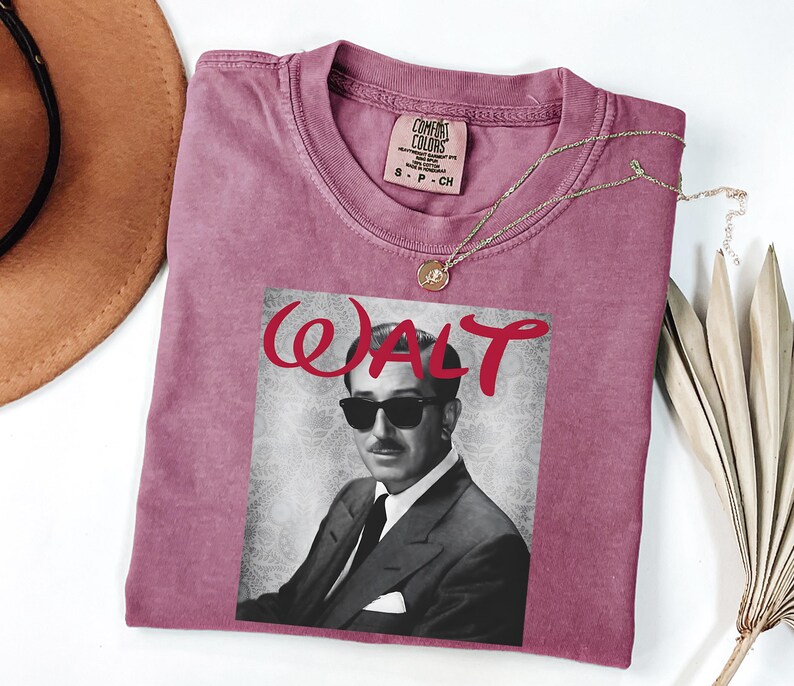 Op de afbeelding: Een stoffig roze t-shirt met een zwart-wit afbeelding van een man met een zonnebril en het woord "WALT" in rode letters. Het shirt heeft een ronde hals en een klein label bij de halslijn. Een gouden ketting met een hanger is over het shirt gedrapeerd.