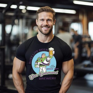 May include: Black t-shirt featuring a cartoon Buzz Lightyear holding a beer mug. The shirt has the text "Buzz Lite Beer" and "Star Command Brewery" with "Best Quality Since 1995" printed on it. The shirt is worn by a person.