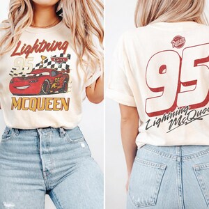 Op de afbeelding: Cr&egrave;mekleurig T-shirt met een afbeelding van de Lightning McQueen raceauto en de tekst "Lightning McQueen 95" op de voorkant. De achterkant van het shirt toont een grote "95" en "Lightning McQueen" in het rood. Het shirt wordt gecombineerd met een blauwe jeans.