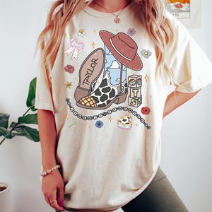 K&ouml;nnte beinhalten: Ein cremefarbenes Kurzarm-T-Shirt mit einem Grafikdruck, der einen Cowboystiefel mit Kuhfleckenmuster, einen braunen Hut, Alphabet-W&uuml;rfel und den Namen TAYLOR zeigt. Das Design enth&auml;lt kleine Sterne, Blumen, Schleifen und Text in einem Kreis.