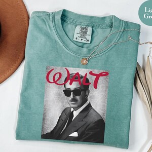 Op de afbeelding: Lichtgroen T-shirt met een zwart-wit portret van een man met een zonnebril, met de naam "WALT" in rode letters. Het shirt heeft een ronde hals en korte mouwen. Een gouden ketting is over het shirt gedrapeerd.