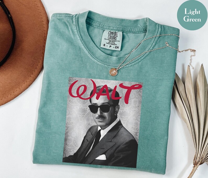 Op de afbeelding: Lichtgroen T-shirt met een zwart-wit portret van een man met een zonnebril, met de naam "WALT" in rode letters. Het shirt heeft een ronde hals en korte mouwen. Een gouden ketting is over het shirt gedrapeerd.