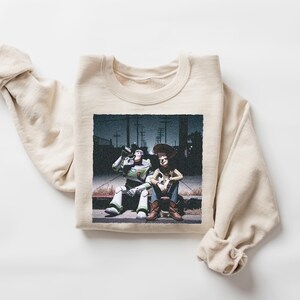 May include: A cream-colored crewneck sweatshirt featuring a graphic print of two animated characters sitting on a curb at night. One character is a space ranger in a white and green suit, and the other is a cowboy in a yellow shirt and brown vest.