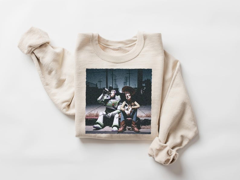 May include: A cream-colored crewneck sweatshirt featuring a graphic print of two animated characters sitting on a curb at night. One character is a space ranger in a white and green suit, and the other is a cowboy in a yellow shirt and brown vest.