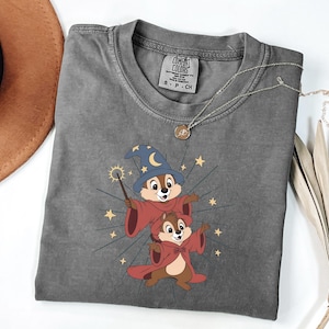May include: A gray Comfort Colors t-shirt featuring a cartoon illustration of two chipmunks dressed as wizards, with a blue hat and red robes. The design includes stars and a magic wand. A silver necklace with a pendant is on the shirt.