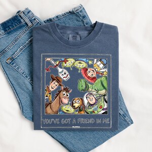 Puede incluir: Una camiseta azul marino con una colorida ilustraci&oacute;n de personajes de la pel&iacute;cula animada "Toy Story". El gr&aacute;fico est&aacute; enmarcado con un borde blanco e incluye el texto "YOU'VE GOT A FRIEND IN ME". La camiseta se combina con unos vaqueros azules.