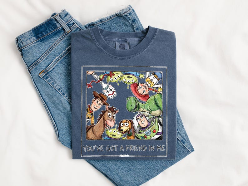 Puede incluir: Una camiseta azul marino con una colorida ilustraci&oacute;n de personajes de la pel&iacute;cula animada "Toy Story". El gr&aacute;fico est&aacute; enmarcado con un borde blanco e incluye el texto "YOU'VE GOT A FRIEND IN ME". La camiseta se combina con unos vaqueros azules.