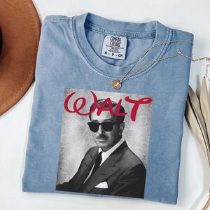 Op de afbeelding: Een lichtblauw Comfort Colors T-shirt met een zwart-wit afbeelding van Walt Disney met een zonnebril. Het woord "WALT" is in rode sierletters boven de afbeelding gedrukt. Een gouden ketting is over het shirt gedrapeerd.
