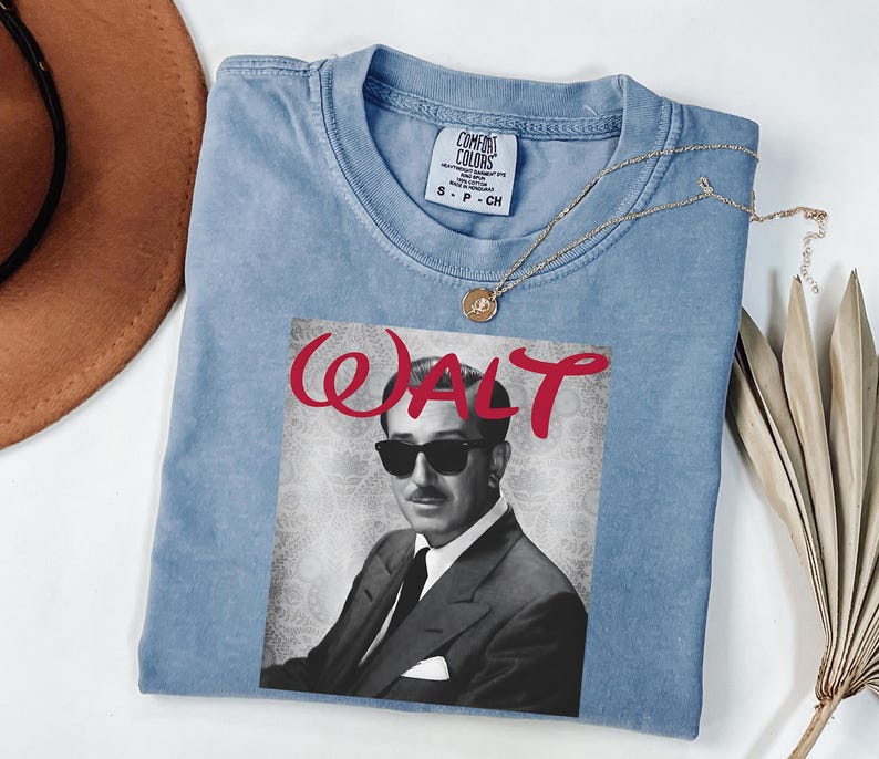 Op de afbeelding: Een lichtblauw Comfort Colors T-shirt met een zwart-wit afbeelding van Walt Disney met een zonnebril. Het woord "WALT" is in rode sierletters boven de afbeelding gedrukt. Een gouden ketting is over het shirt gedrapeerd.