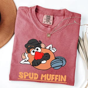 Puede incluir: Una camiseta Comfort Colors color granate con un dise&ntilde;o de dibujos animados de Mr. Potato Head, que incluye un sombrero negro, una nariz roja y una bola de lana gris. El texto naranja dice "SPUD MUFFIN". Un collar dorado est&aacute; sobre la camiseta.
