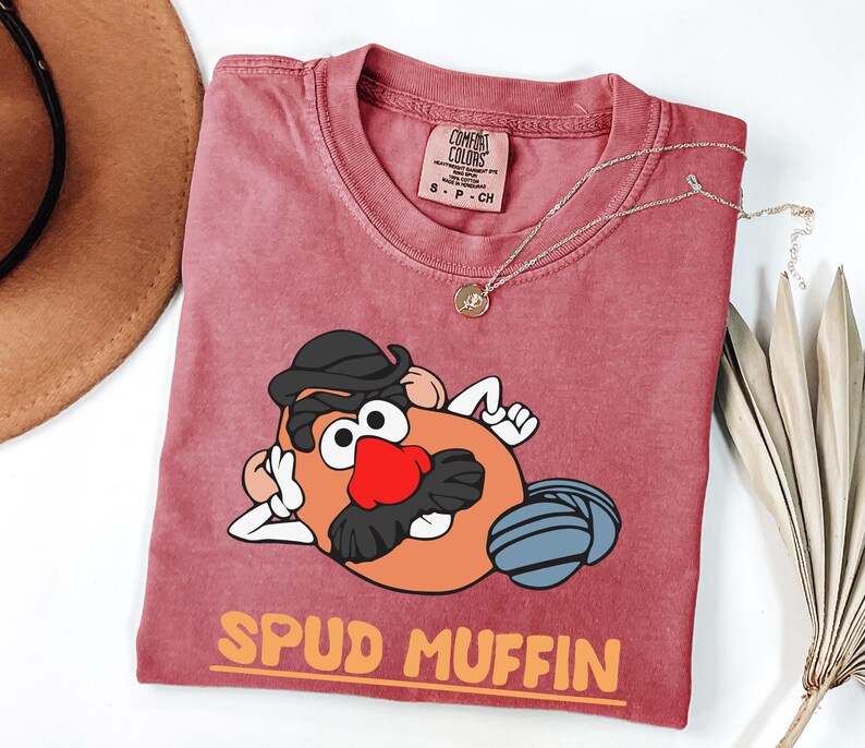 Puede incluir: Una camiseta Comfort Colors color granate con un dise&ntilde;o de dibujos animados de Mr. Potato Head, que incluye un sombrero negro, una nariz roja y una bola de lana gris. El texto naranja dice "SPUD MUFFIN". Un collar dorado est&aacute; sobre la camiseta.
