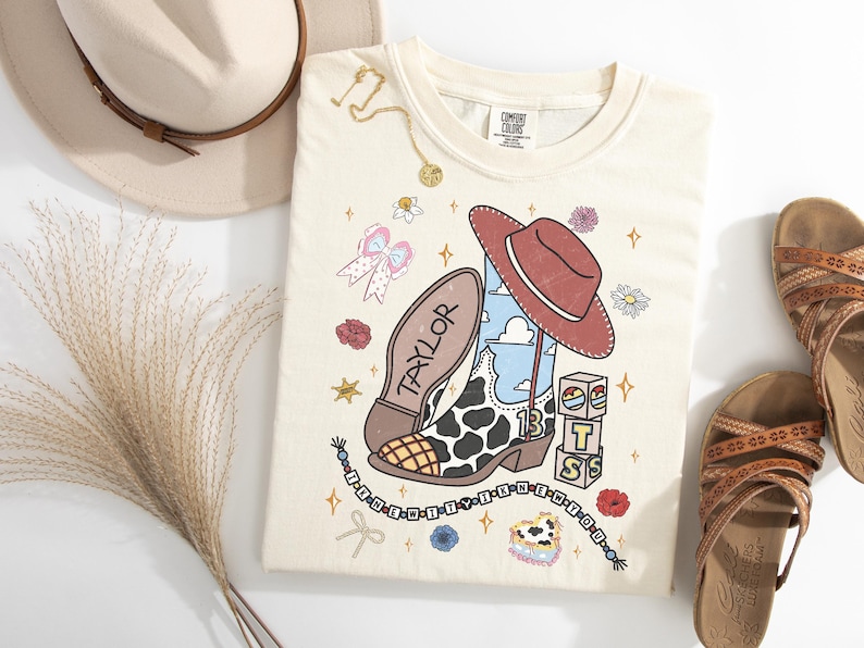 Puede incluir: Una camiseta de color crema con un estampado gr&aacute;fico de una bota vaquera con manchas de vaca, un sombrero rojo, bloques de letras que forman TS y el nombre Taylor. El dise&ntilde;o incluye peque&ntilde;as ilustraciones de flores, lazos y una cadena.