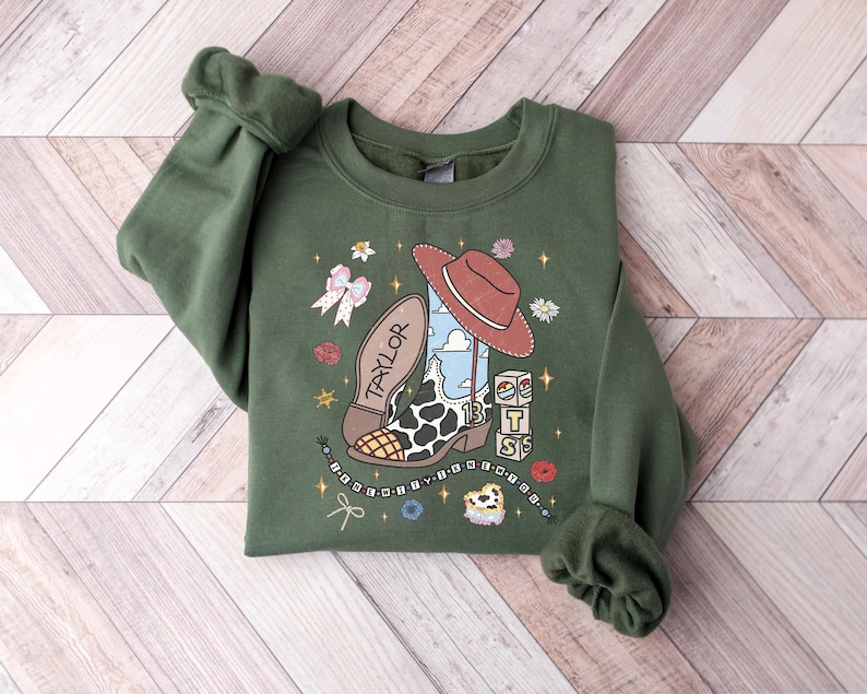 K&ouml;nnte beinhalten: Ein olivgr&uuml;ner Rundhals-Pullover mit einem Grafikdruck, der einen Cowboystiefel mit Kuhfleckenmuster, einen roten Cowboyhut, eine Schleife, W&uuml;rfel mit den Buchstaben BTS und ein Perlenarmband zeigt. Die Stiefelsohle ist mit dem Namen TAYLOR beschriftet.