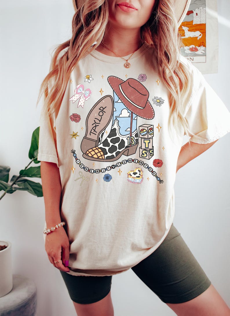 Puede incluir: Una camiseta de manga corta color crema con un estampado gr&aacute;fico de una bota vaquera con patr&oacute;n de manchas de vaca, un sombrero marr&oacute;n, bloques de letras y el nombre TAYLOR. El dise&ntilde;o incluye peque&ntilde;as estrellas, flores, lazos y texto.