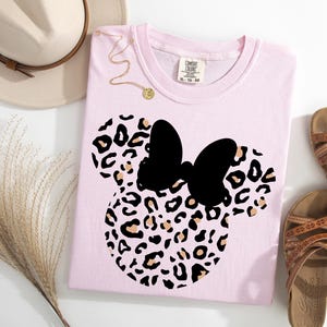 May include: A light pink t-shirt featuring a black bow and a leopard print design in the shape of a mouse. The shirt is next to a beige hat, a gold necklace, and brown sandals.