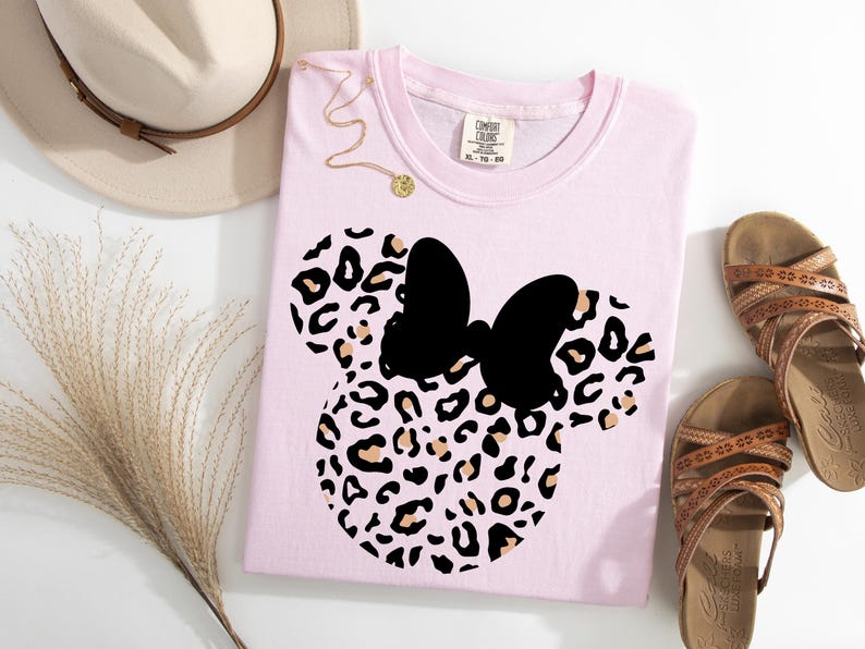 May include: A light pink t-shirt featuring a black bow and a leopard print design in the shape of a mouse. The shirt is next to a beige hat, a gold necklace, and brown sandals.