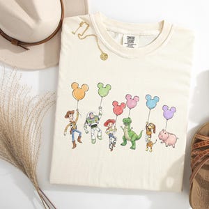 May include: A cream-coloured t-shirt featuring a graphic print of six animated characters holding colourful Mickey Mouse-shaped balloons. The characters include a cowboy, a space ranger, a cowgirl, a green dinosaur, a dachshund, and a pink pig.