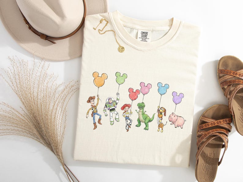 May include: A cream-coloured t-shirt featuring a graphic print of six animated characters holding colourful Mickey Mouse-shaped balloons. The characters include a cowboy, a space ranger, a cowgirl, a green dinosaur, a dachshund, and a pink pig.