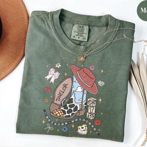 Puede incluir: Una camiseta verde musgo con un estampado gr&aacute;fico de una bota vaquera con patr&oacute;n de manchas de vaca, un sombrero rojo, el nombre Taylor, el n&uacute;mero 13 y bloques apilados. El dise&ntilde;o incluye peque&ntilde;as flores, un lazo y un collar de cuentas.