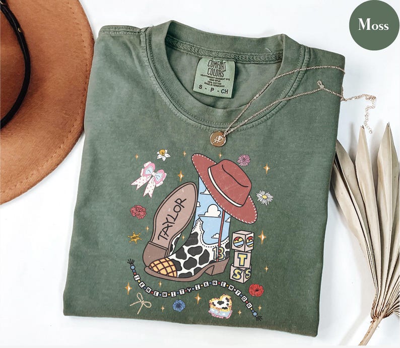 Puede incluir: Una camiseta verde musgo con un estampado gr&aacute;fico de una bota vaquera con patr&oacute;n de manchas de vaca, un sombrero rojo, el nombre Taylor, el n&uacute;mero 13 y bloques apilados. El dise&ntilde;o incluye peque&ntilde;as flores, un lazo y un collar de cuentas.