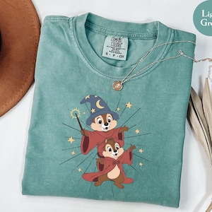 May include: Light green t-shirt featuring a graphic of two cartoon chipmunks dressed as wizards, with a wizard hat and red robes. The shirt has a crew neck and a Comfort Colors tag. A gold necklace is draped across the shirt.