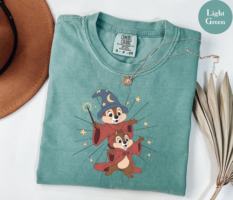 May include: Light green t-shirt featuring a graphic of two cartoon chipmunks dressed as wizards, with a wizard hat and red robes. The shirt has a crew neck and a Comfort Colors tag. A gold necklace is draped across the shirt.