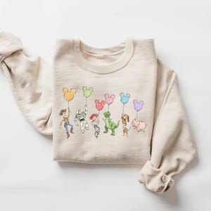 Puede incluir: Una sudadera de cuello redondo en color beige con una ilustraci&oacute;n gr&aacute;fica colorida de varios personajes de juguetes animados, cada uno sosteniendo un globo de color pastel con forma de cabeza de rat&oacute;n. El dise&ntilde;o incluye a Woody, Buzz Lightyear, Jessie, Rex, Slinky Dog y Hamm.