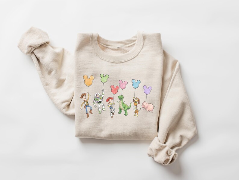 Puede incluir: Una sudadera de cuello redondo en color beige con una ilustraci&oacute;n gr&aacute;fica colorida de varios personajes de juguetes animados, cada uno sosteniendo un globo de color pastel con forma de cabeza de rat&oacute;n. El dise&ntilde;o incluye a Woody, Buzz Lightyear, Jessie, Rex, Slinky Dog y Hamm.