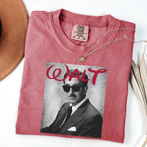 Op de afbeelding: Een rood Comfort Colors T-shirt met een zwart-wit afbeelding van Walt Disney met een zonnebril. Het woord "WALT" is in rode cursieve letters boven de afbeelding gedrukt. Een gouden ketting met een hanger ligt op het shirt.