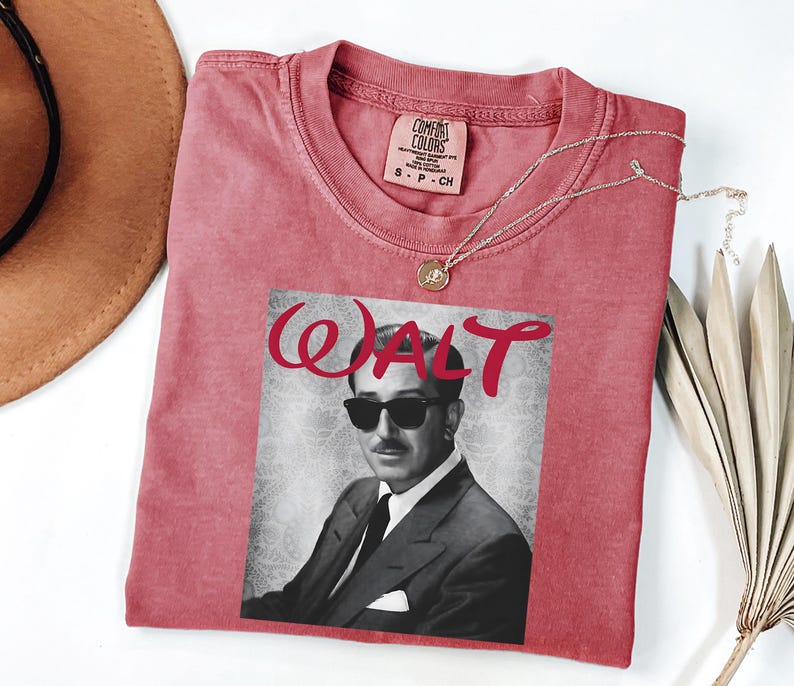 Op de afbeelding: Een rood Comfort Colors T-shirt met een zwart-wit afbeelding van Walt Disney met een zonnebril. Het woord "WALT" is in rode cursieve letters boven de afbeelding gedrukt. Een gouden ketting met een hanger ligt op het shirt.