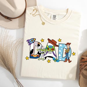 May include: Cream-coloured t-shirt featuring the word "DAD" in colourful, cartoon-style letters. The letters are decorated with images of characters from a popular animated film. Yellow stars are scattered around the letters.
