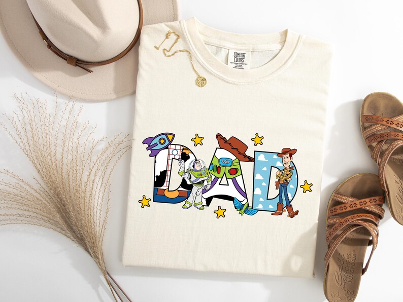 May include: Cream-coloured t-shirt featuring the word "DAD" in colourful, cartoon-style letters. The letters are decorated with images of characters from a popular animated film. Yellow stars are scattered around the letters.