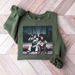 May include: An olive green crewneck sweatshirt featuring a graphic print of the characters Buzz Lightyear and Woody sitting together on a curb at night. The design shows the two figures in a dark outdoor setting with utility poles in the background.