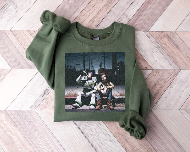 May include: An olive green crewneck sweatshirt featuring a graphic print of the characters Buzz Lightyear and Woody sitting together on a curb at night. The design shows the two figures in a dark outdoor setting with utility poles in the background.