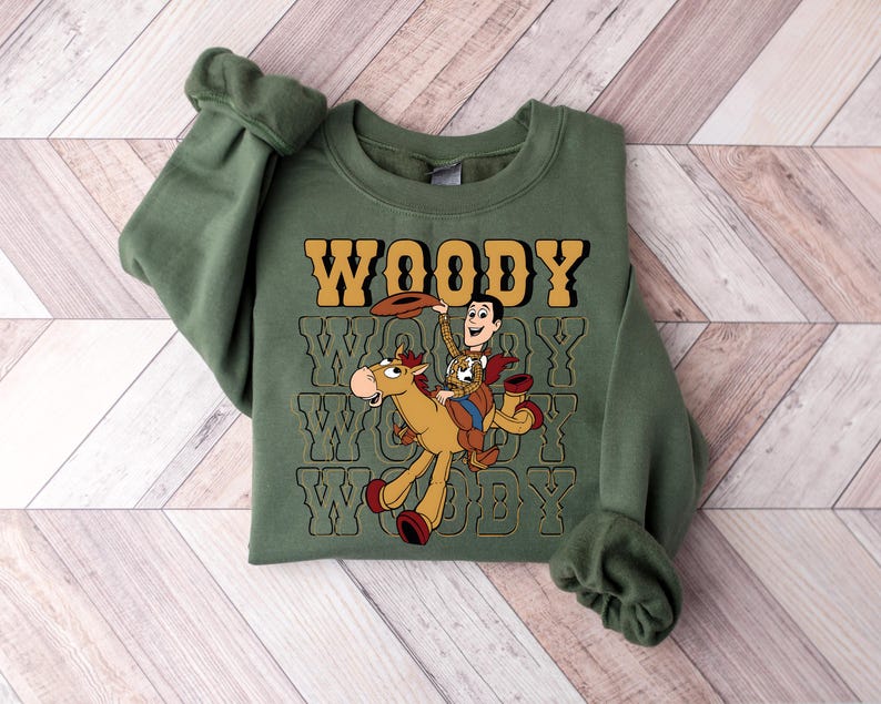 May include: An olive green sweatshirt featuring a graphic of Woody from Toy Story riding a horse. The word "WOODY" is printed in a retro font above the graphic, repeated below. The sweatshirt has long sleeves and a crew neck.