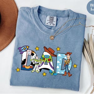 May include: Blue jean coloured t-shirt with the word "DAD" in colourful Toy Story-themed letters. The "D" features a rocket ship, the "A" has Buzz Lightyear, and the other "D" has Woody. The shirt has a crew neck and a small tag.