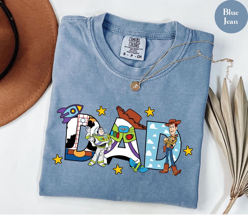 May include: Blue jean coloured t-shirt with the word "DAD" in colourful Toy Story-themed letters. The "D" features a rocket ship, the "A" has Buzz Lightyear, and the other "D" has Woody. The shirt has a crew neck and a small tag.