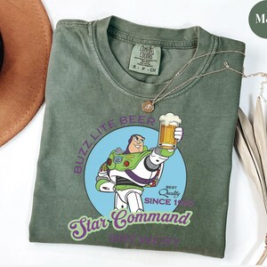 May include: Moss green t-shirt featuring a graphic of Buzz Lightyear holding a beer, with the text "Buzz Lite Beer" and "Star Command". The shirt has a crew neck and short sleeves. A gold necklace is draped across the shirt.