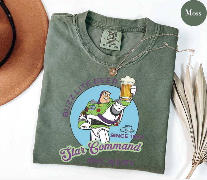 May include: Moss green t-shirt featuring a graphic of Buzz Lightyear holding a beer, with the text "Buzz Lite Beer" and "Star Command". The shirt has a crew neck and short sleeves. A gold necklace is draped across the shirt.