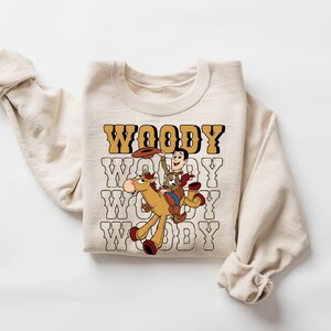 May include: Cream-colored sweatshirt featuring a graphic of Woody from Toy Story riding a horse. The word "WOODY" is printed in a retro font above and below the graphic. The sweatshirt has long sleeves.