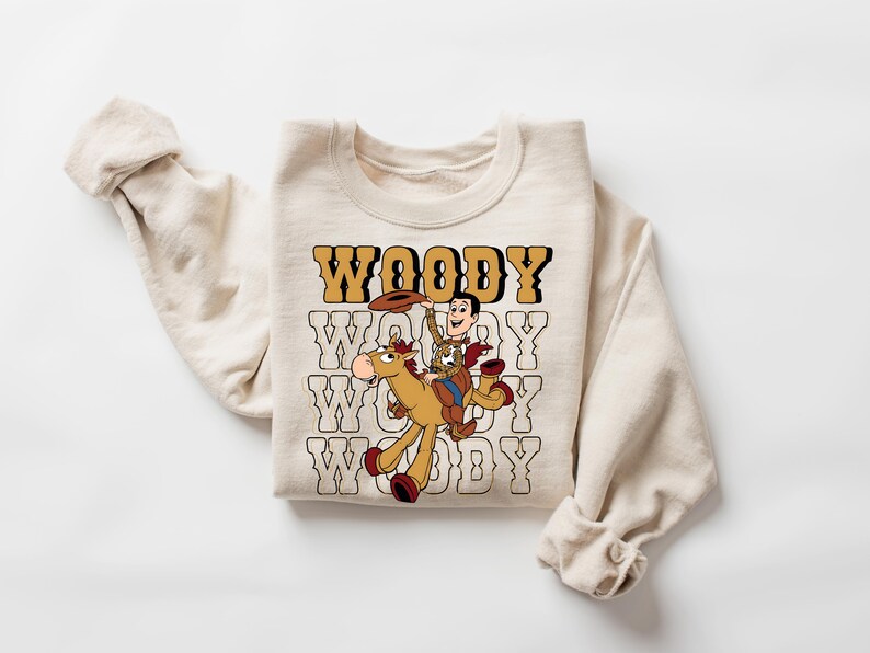 May include: Cream-colored sweatshirt featuring a graphic of Woody from Toy Story riding a horse. The word "WOODY" is printed in a retro font above and below the graphic. The sweatshirt has long sleeves.