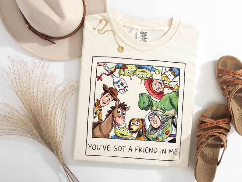 Puede incluir: Camiseta color crema con una ilustraci&oacute;n colorida de personajes de Toy Story, enmarcada en un borde negro. El texto "YOU'VE GOT A FRIEND IN ME" est&aacute; impreso debajo. La imagen se complementa con sandalias y un sombrero.