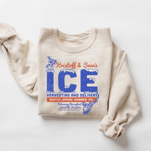 Puede incluir: Una sudadera color crema con mangas enrolladas. La parte delantera presenta un gr&aacute;fico azul y rojo que dice "Kristoff & Sven's ICE" con el texto "HARVESTING AND DELIVERY" y "WINTER SPRING SUMMER FALL". Tambi&eacute;n se ven un mu&ntilde;eco de nieve y un reno de dibujos animados.